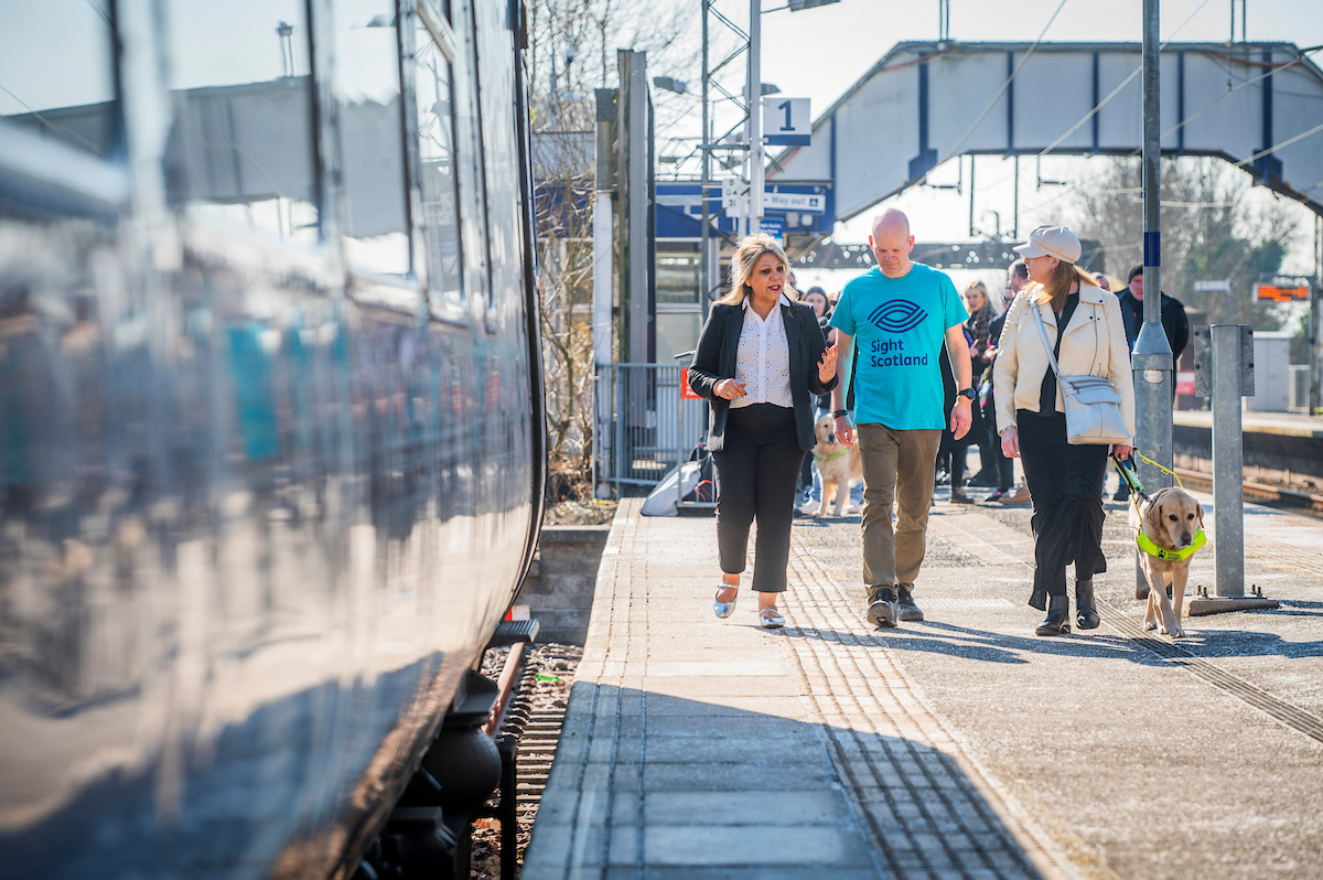 Equalities Minister Kaukab Stewart walks down a train platform with Craig Spalding, Chief Executive of Sight Scotland and Sight Scotland Veteran, and a visually impaired passenger who is with their guide dog.