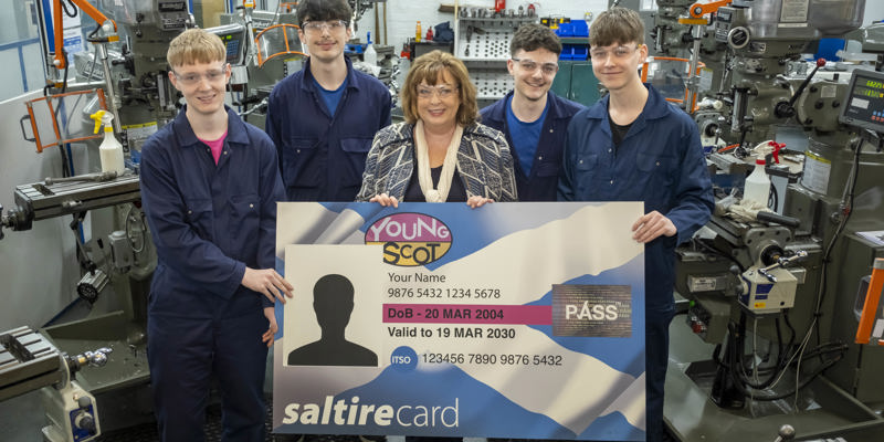 Transport Secretary Fiona Hyslop poses for a photo with a group of young apprentices in a mechanics workshop. The apprentices are wearing overalls and protective glasses. Together, they are holding up a giant version of a young persons' bus pass card.