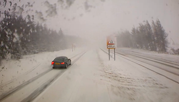 From the view through the windscreen of a gritter, a car drives along a road in dangerous, snowy conditions.