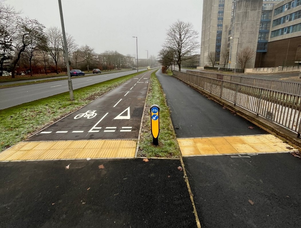 A newly constructed segregated cycling lane with road markings and tactile paving beside a main road, running parallel to a footpath separated by a narrow verge. A small blue wayfinding bollard stands between the cycle lane and the footpath.
