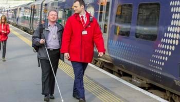A rail worker helps guide a blind passenger down a station platform.