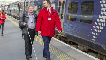 A rail worker helps guide a blind passenger down a station platform.