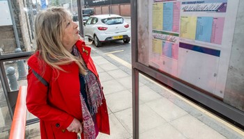 A woman using a walking stick examines timetables at a bus stop.