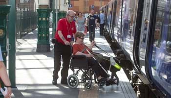 A rail worker assists a wheelchair user and their guide dog on to a train.