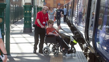 A rail worker assists a wheelchair user and their guide dog on to a train.