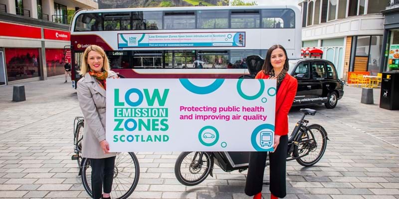 Transport Minister Jenny Gilruth and Environment Minister Màiri McAllan celebrate the launch of Low Emission Zones in Scotland. They are standing on a street in Edinburgh, holding a sign that says &#x27;Low Emission Zones Scotland. Protecting public health and improving air quality&#x27;. In the background there is a bus, taxi and two bikes. Edinburgh Castle is visible in the distance.