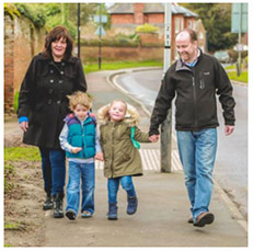 2 parents and 2 young children holding hands and walking along a pavement with a pedestrian crossing in the background.