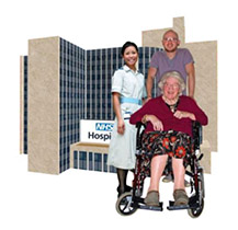 An older woman in a wheelchair with 2 NHS staff beside her. Behind them is an NHS hospital building.