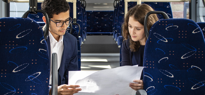 2 people sitting on a bus. They're holding a large piece of paper between them that they are reading.