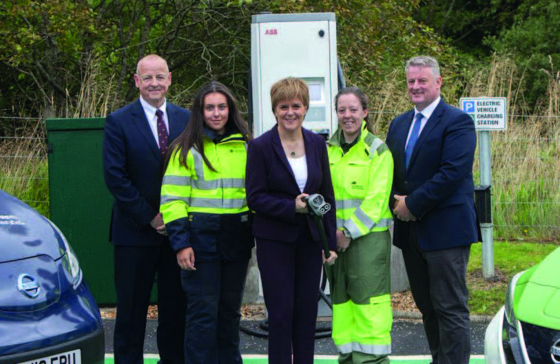 The First Minister Nicola Sturgeon has 2 people at each side. They are standing in front of an electric vehicle charging point and the First Minister is holding the charger.