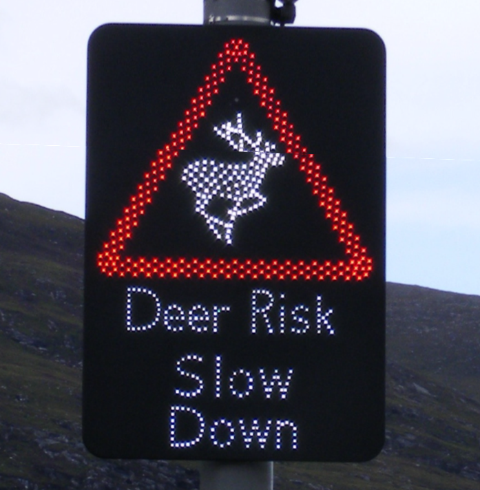 Dot matrix red triangle warning sign with silhouette of deer, with text saying "Deer Risk Slow Down"