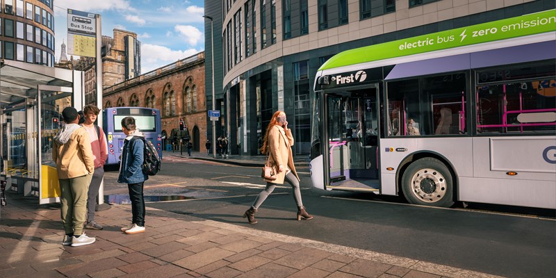 A sunny day on a street in Glasgow. A group of young people wait at a bus stop. One young woman steps out to the road to board a bus which has stopped.