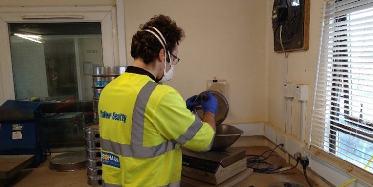 William, wearing PPE and a face mask, grading material using accurate weighing equipment in a laboratory
