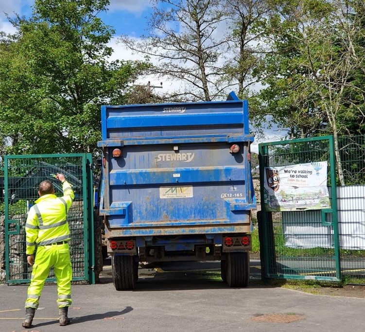 A man in protective clothing directs a lorry which is backing through a narrow gateway