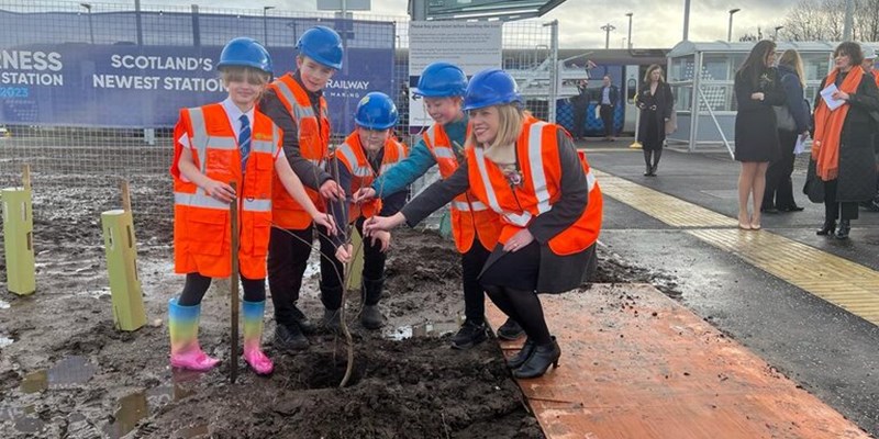Transport Minister Jenny Gilruth poses for a photo with a group of children at the official opening of Inverness Airport rail station. They are wearing hard hats and hi vis jackets while planting a tree.
