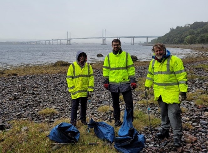 A photograph of 3 Jacobs employees in high vis jackets standing on North Kessock beach with trashbags and litter pickers