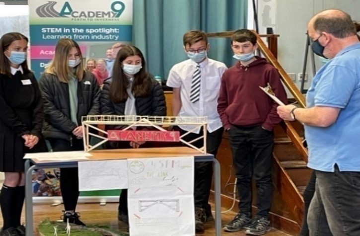 Photograph of five school pupils standing with their constructed bridge for the bridge competition. Man is scoring their submission