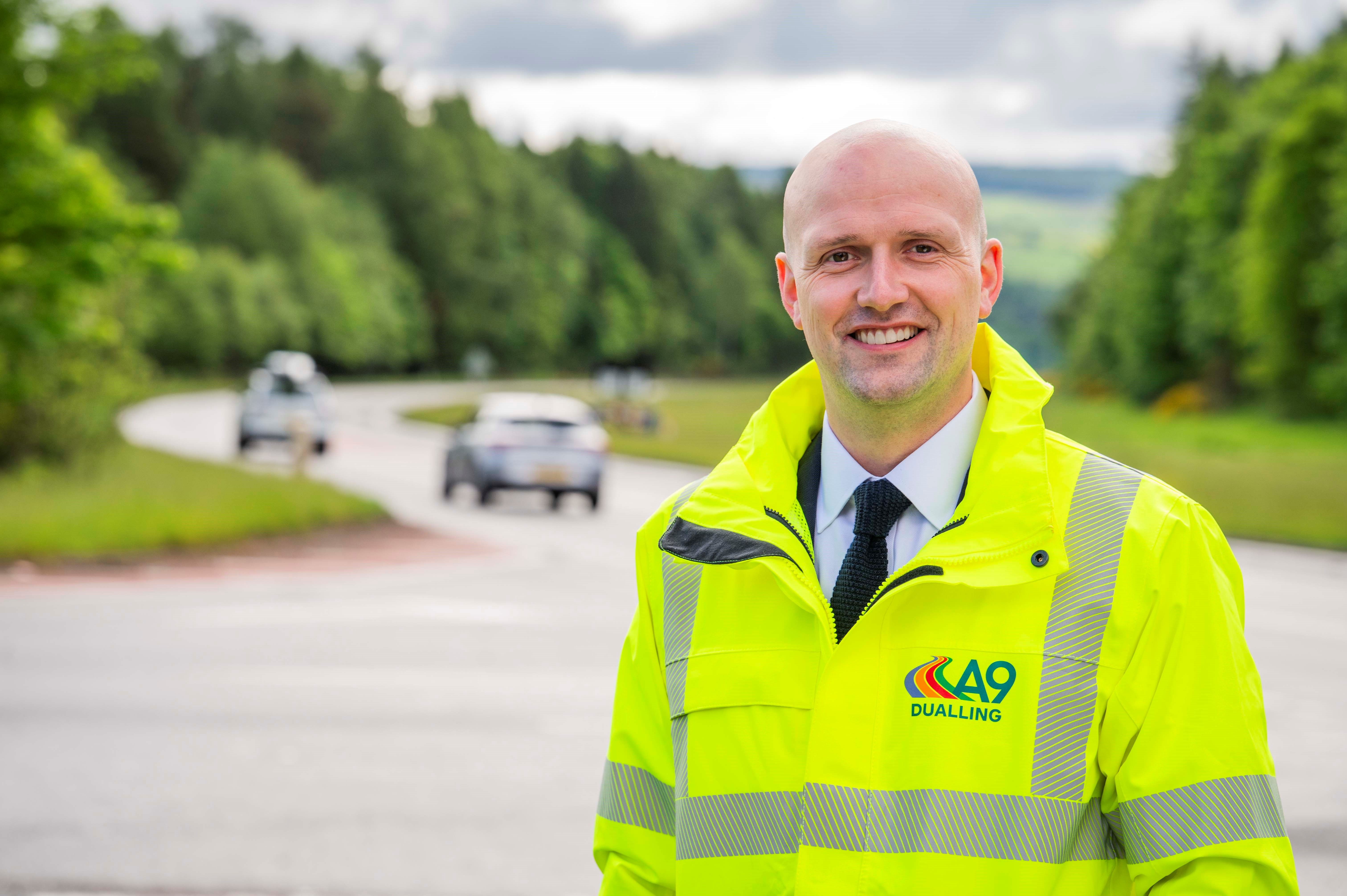 Transport Secretary Stephen Flynn smiles for the camera at the side of the A9. He is wearing hi-vis A9 Dualling branded clothing.