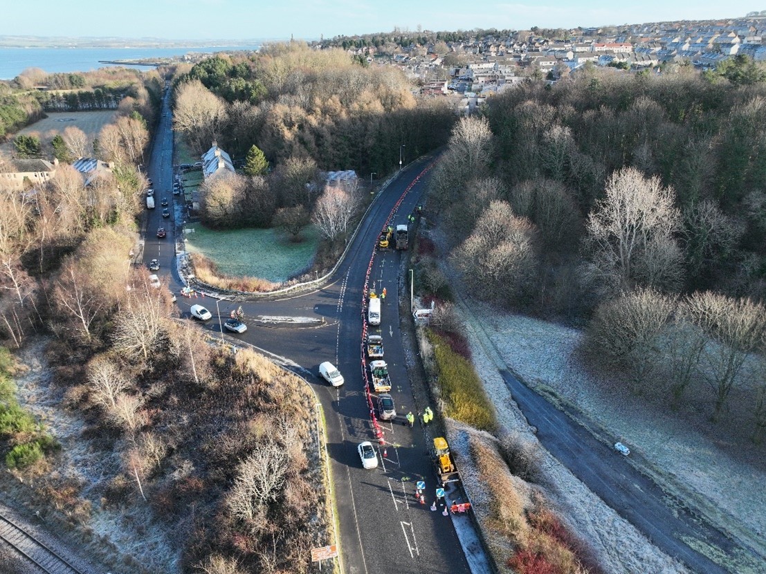 An aerial view of a road where construction vehicles and workers are carrying out road improvement works, with traffic cones and lane closures guiding cars through the area.