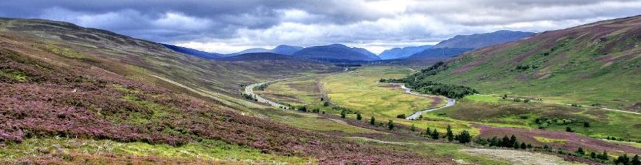 Panoramic landscape photograph showing a broad Highland valley surrounded by rolling mountains under a cloudy sky. Purple heather covers foreground slopes, while a curving road, a railway, a meandering river, green fields, and scattered trees lead toward distant blue peaks.