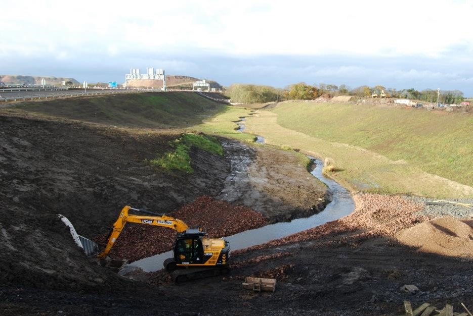 Photograph showing an excavator reshaping a muddy stream channel within a broad graded construction site. Exposed soil, rock piles, drainage water, grassy embankments, and construction compound mark ongoing watercourse realignment work.