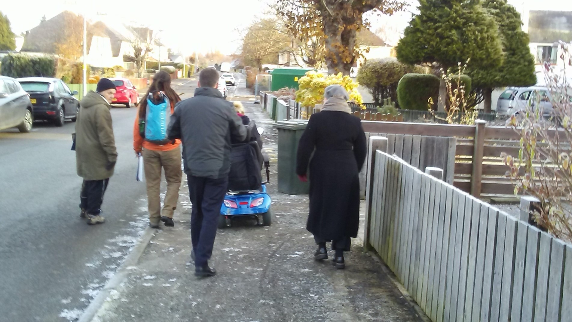 A person in a mobility scooter, a local councillor, a road safety officer, a regional transport partnership officer and a Living Streets officer walk along a street in a residential area.