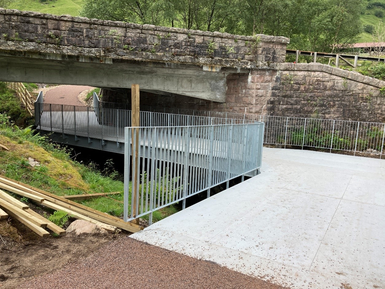 In a rural setting, a new pedestrian bridge crossing a river, under a road bridge.