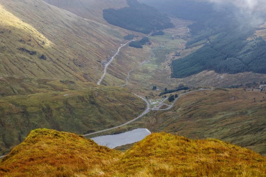 Photograph showing a mountainous Scottish valley viewed from an elevated hillside. A winding road, narrow stream, small loch, scattered buildings, forested slopes, and golden-green moorland define rugged landscape.