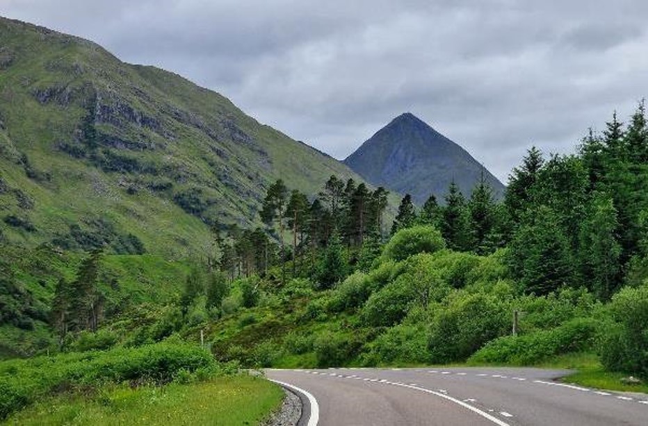 A Scottish road with lush vegetation, variety of trees and rugged mountains in the background.