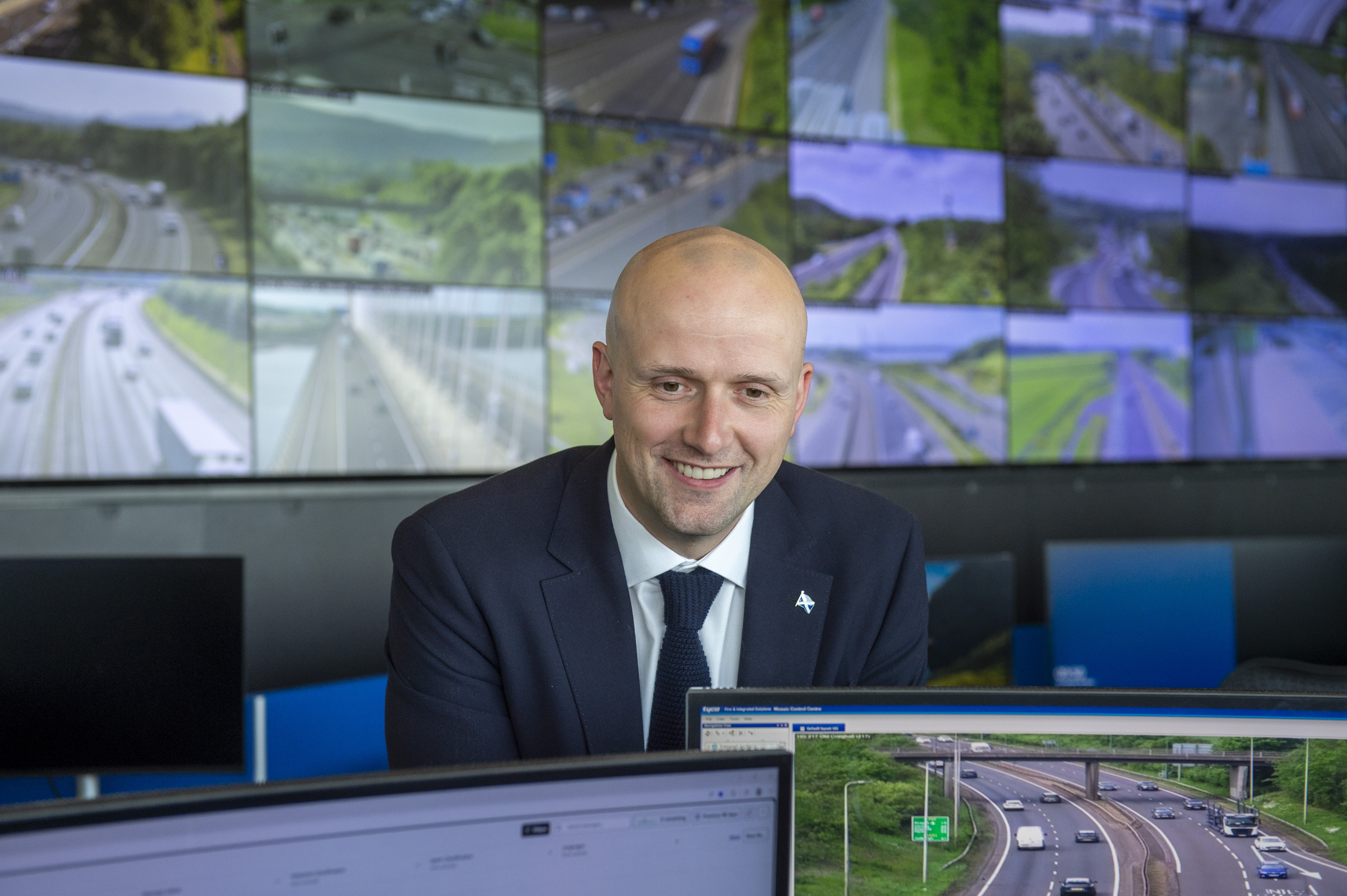 Transport Secretary Stephen Flynn smiles for a photo at the Traffic Scotland National Control Centre. Many monitors in the background show live footage from Scotland's trunk road network.