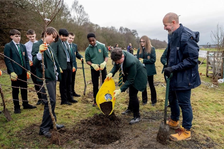 Photograph showing school pupils involved in conservation activities. The pupils are wearing green school uniforms and planting a young tree outdoors alongside an adult environmental volunteer. One pupil holds the tree while another tips compost from a yellow sack. There is a canal in the background.