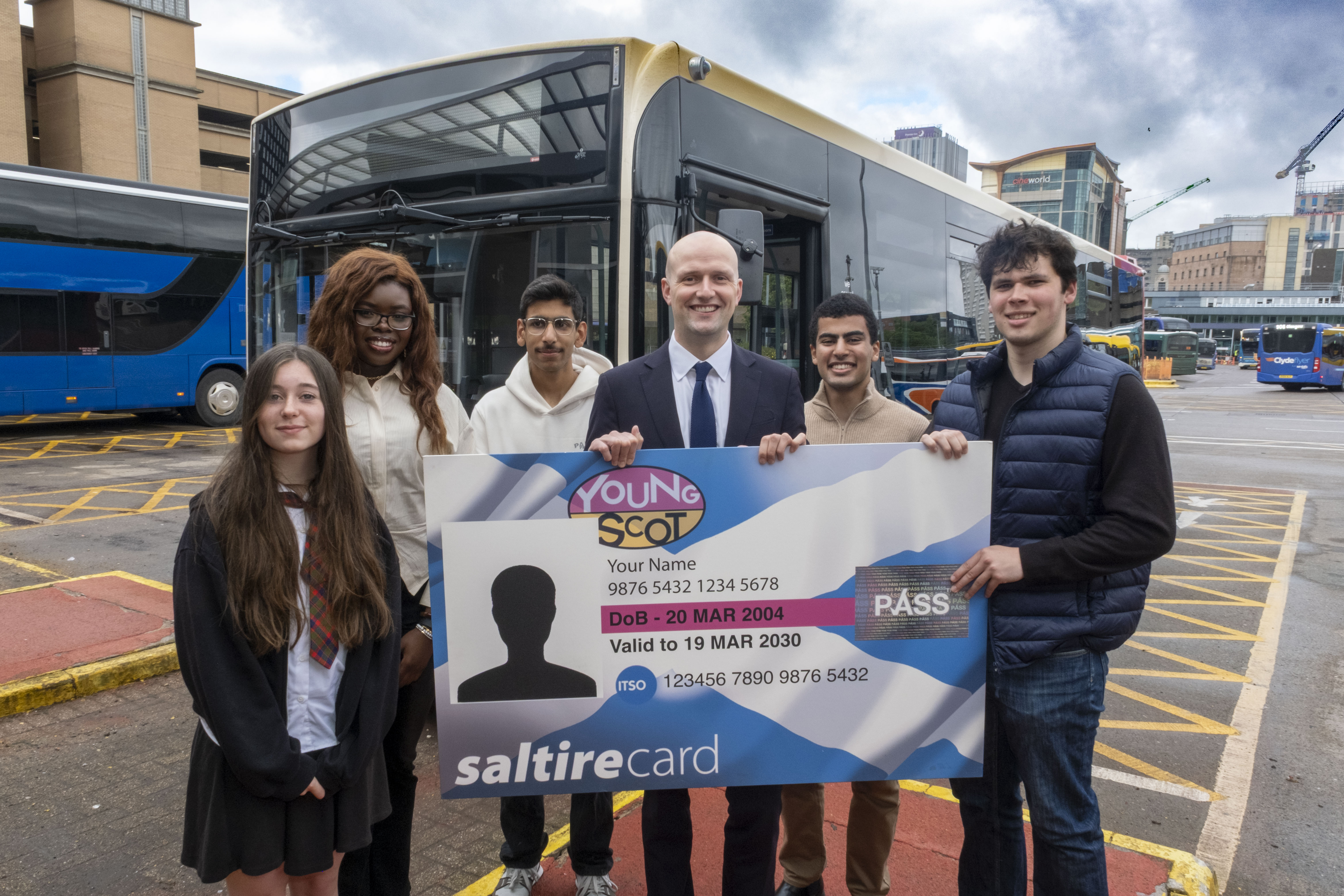 Transport Secretary Stephen Flynn  poses for a photo with a group of young people under 22. They are at a bus station, with a bus behind them. Together, they hold up a giant Young Scot National Entitlement Card.