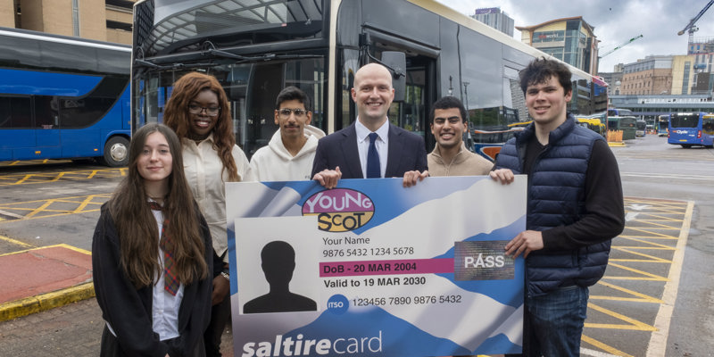 Transport Secretary Stephen Flynn  poses for a photo with a group of young people under 22. They are at a bus station, with a bus behind them. Together, they hold up a giant Young Scot National Entitlement Card.