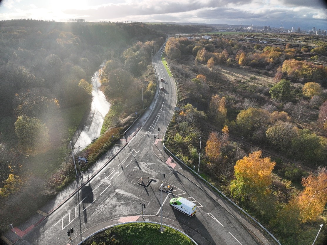 Aerial photo of a junction on a rural road showing new road safety interventions such as refreshed lane markings, a central traffic island, traffic signals, and newly defined pedestrian crossing points bordered by green space.