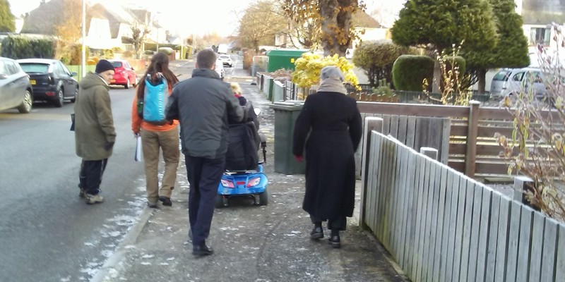 A person in a mobility scooter, a local councillor, a road safety officer, a regional transport partnership officer and a Living Streets officer walk along a street in a residential area.